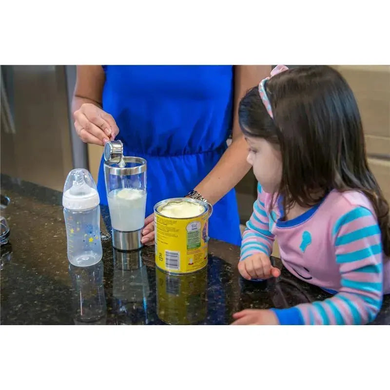 A woman in a blue dress uses a measuring spoon to add formula to a stainless steel mixer cup, while a young girl watches. A baby bottle and a can of formula are on the counter.