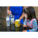 A woman in a blue dress uses a measuring spoon to add formula to a stainless steel mixer cup, while a young girl watches. A baby bottle and a can of formula are on the counter.
