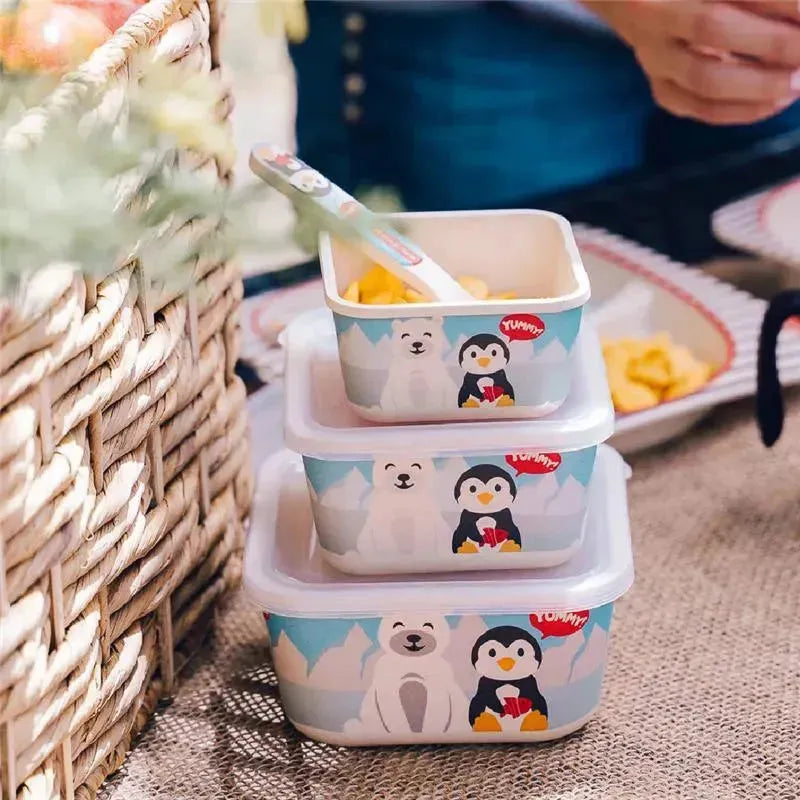 Three bamboo food containers featuring penguin and polar bear designs, placed on a table with snacks and a woven basket.