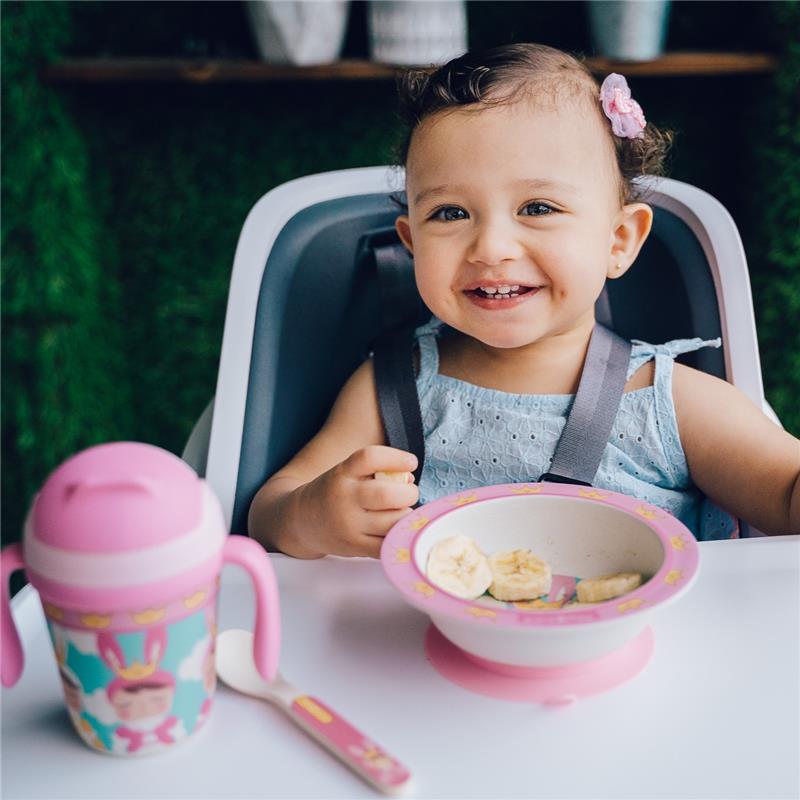 Smiling toddler in a high chair holding a spoon, with a divided plate of bananas and a pink sippy cup beside her.