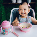 Smiling toddler in a high chair holding a spoon, with a divided plate of bananas and a pink sippy cup beside her.