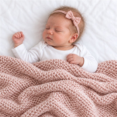 A light pink knit blanket drapes over a sleeping baby with a pink bow headband, resting on a white surface.