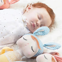 Sleeping baby girl with red hair in white dress cuddling a blue polka dot plush doll and surrounded by other dolls.