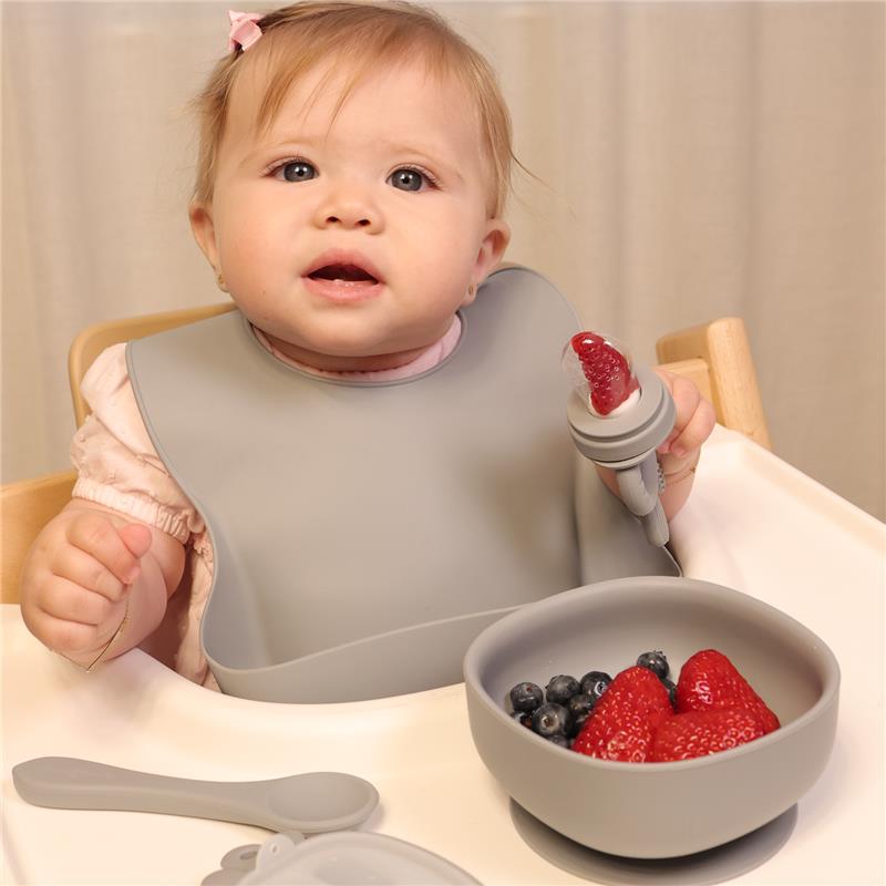 Toddler wearing a gray silicone bib, holding a utensil with fruit in front of a bowl of strawberries and blueberries.