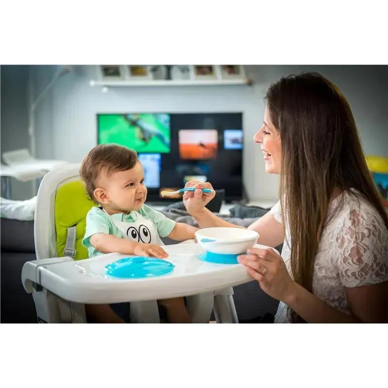 Mother feeding a smiling baby in a high chair, with a silicone baby bib, a bowl, and a spoon visible.