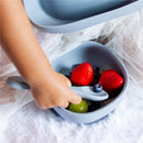 A child’s hand reaching into a dusty blue suction bowl filled with strawberries, blueberries, and grapes, with a spoon nearby.