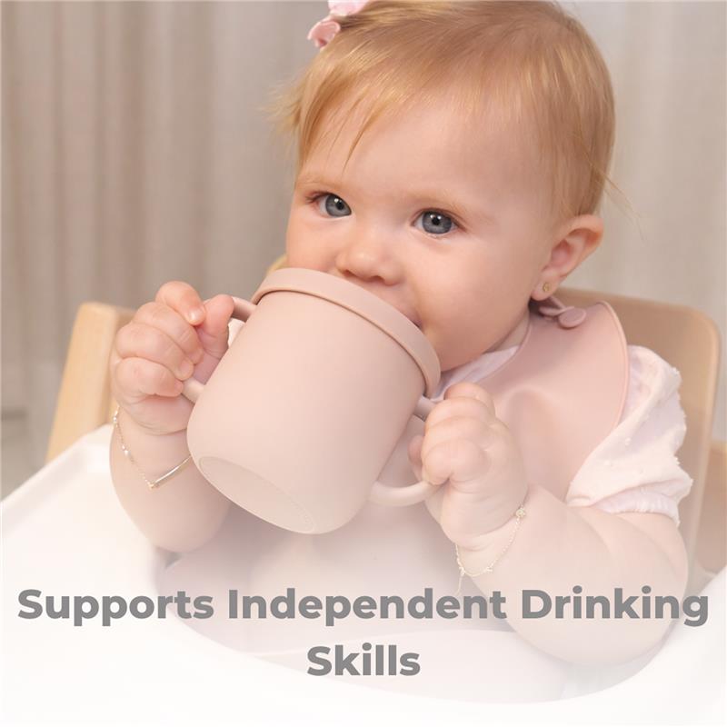 A baby with light hair holds a rose pink training cup, promoting independent drinking skills.