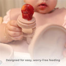 Baby holds a silicone feeder with fruit puree, featuring a rose pink training cup and suction bowl in the background.