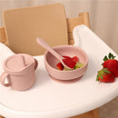 Rose pink silicone feeding set includes a cup, bowl with spoon, and fresh strawberries on a high chair tray.