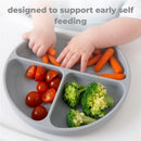Baby hands reaching for carrot sticks and cherry tomatoes in a grey suction plate, designed for early self-feeding.