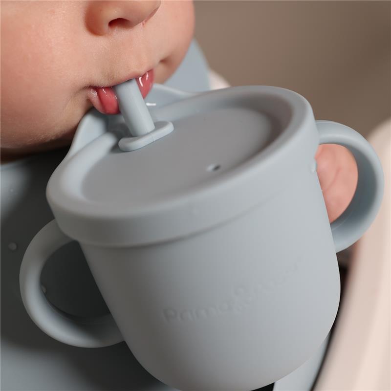 Training cup for toddlers in soft grey silicone, featuring a straw and easy-grip handles, being held by a child.