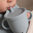 Training cup for toddlers in soft grey silicone, featuring a straw and easy-grip handles, being held by a child.