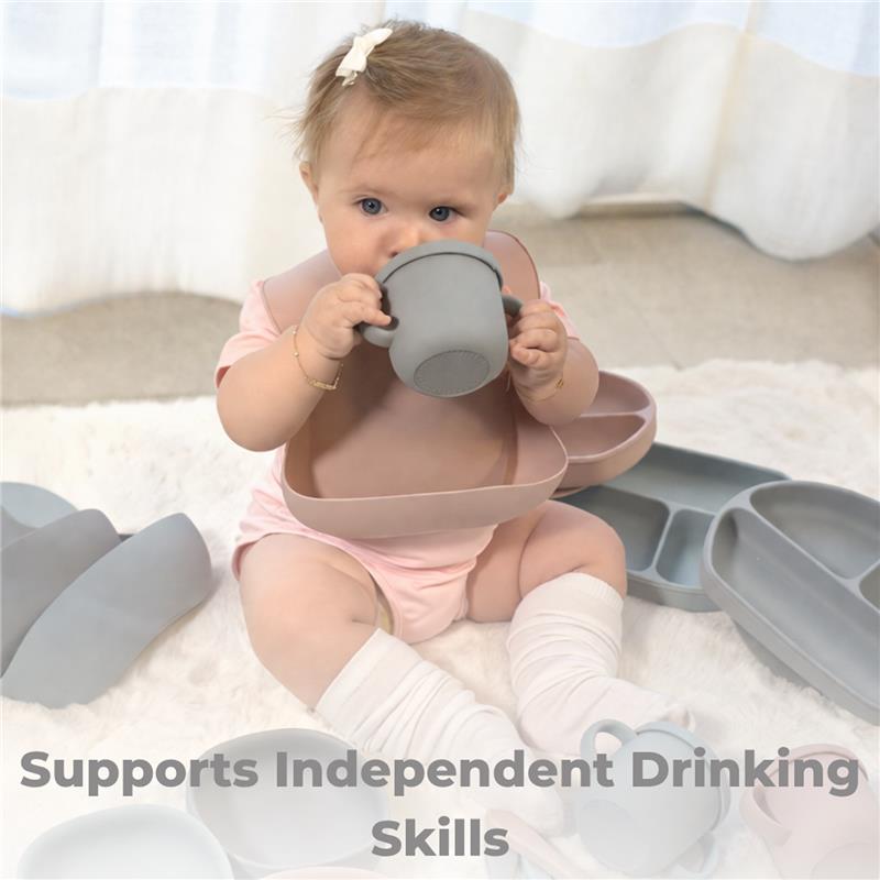 Baby wearing a pink shirt holds a grey training cup, surrounded by silicone feeding tools, promoting independent drinking skills.