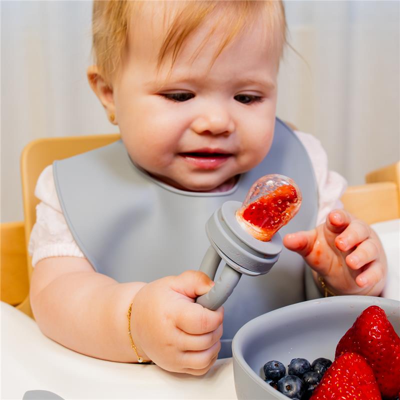 Baby enjoying food from a silicone feeder, holding it in one hand, with a bowl of strawberries and blueberries in front.