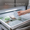 Silicone carrot-shaped freezer tray for storing baby food, held by a hand in a refrigerator, alongside sealed food containers.