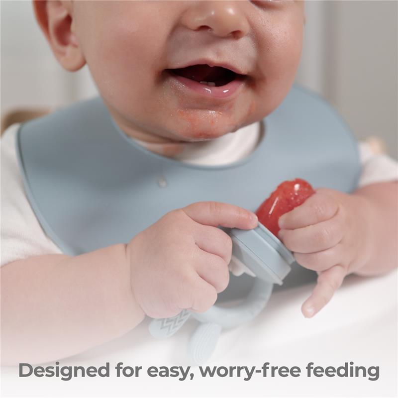 Baby holding a Primo Passi Silicone Food Feeder in rose pink, enjoying a piece of fruit, while wearing a light blue bib.