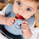 Baby using a Primo Passi Silicone Food Feeder, holding the feeder with fruit inside, wearing a blue bib.