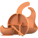 Silicone baby feeding set by Quark, including a divided plate, bowl, bib, spoon, and fork in a warm orange color.