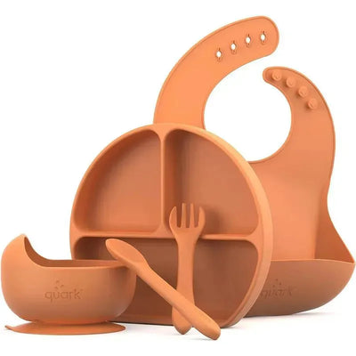 Silicone baby feeding set by Quark, including a divided plate, bowl, bib, spoon, and fork in a warm orange color.