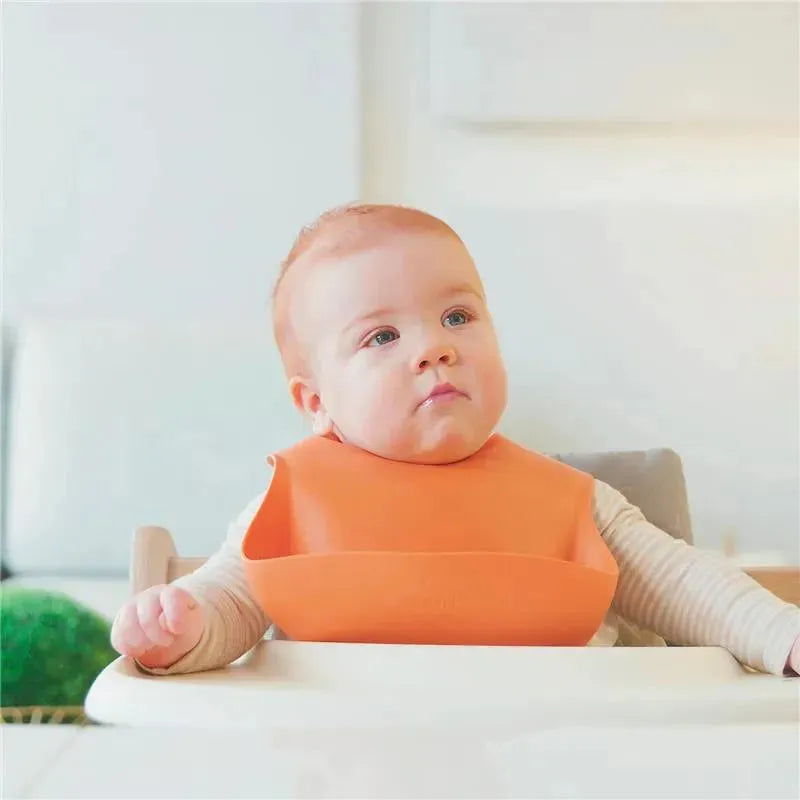 Baby seated in a high chair wearing an orange silicone bib, looking thoughtfully to the side in a bright, airy space.