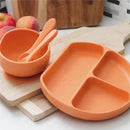 Silicone baby feeding set featuring an orange divided plate, bowl, and matching utensils, arranged on a wooden surface.