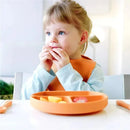 A child in a bib sits at a table, holding fruit slices from a peach-colored silicone feeding plate.