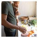 A parent holds a baby while preparing food with a Quark Quook 5-In-1 Baby Food Processor, surrounded by fresh ingredients.