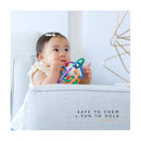 Baby holding a colorful Quark Thiingy teething toy, looking curiously while seated on a light gray couch.