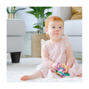 Baby girl in a pink dress sits on a soft rug, holding a colorful Quark Thiingy teething toy with various rings.