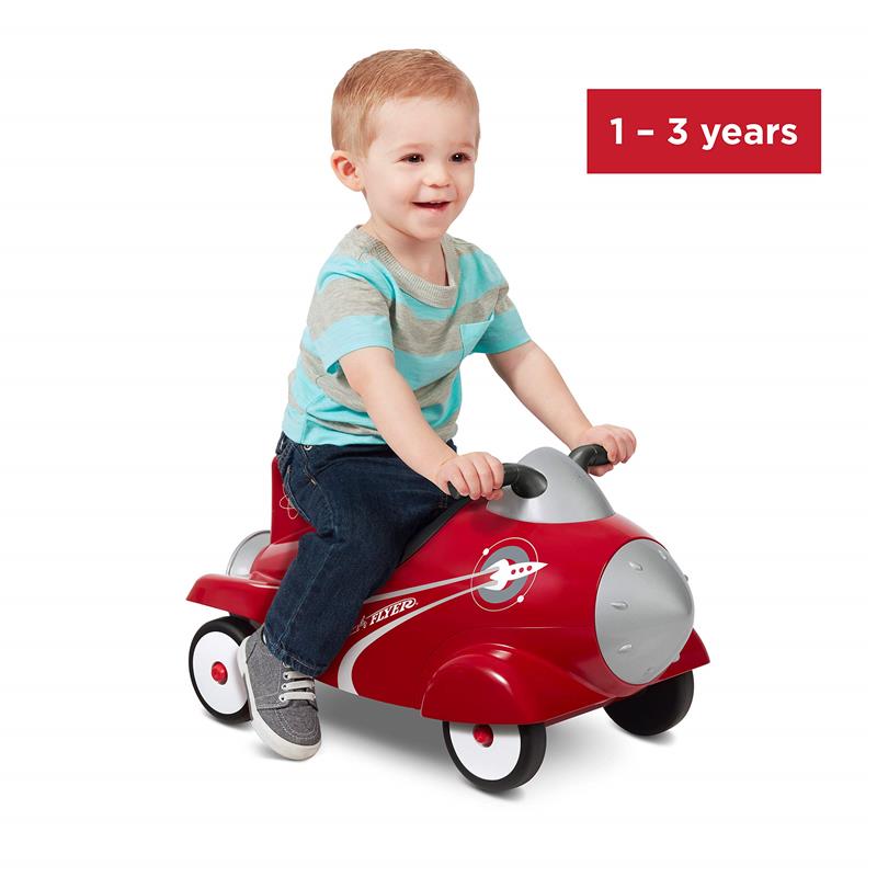 A toddler sits on a red Radio Flyer Retro Rocket Ride-On toy car, smiling while holding the steering wheel.