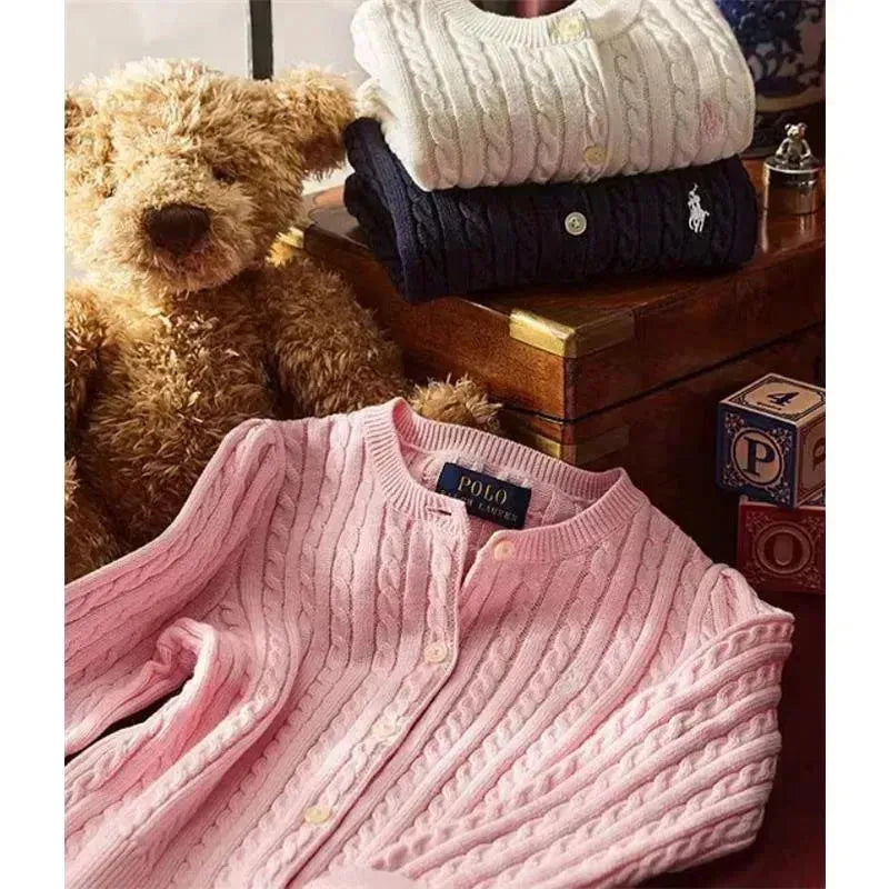 Ralph Lauren Baby pink long-sleeve cashmere cable knit cardigan sweater displayed in front of a teddy bear and stacked sweaters.