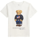White short-sleeve t-shirt featuring a playful bear in a navy sweater with an American flag, styled with jeans and shoes.