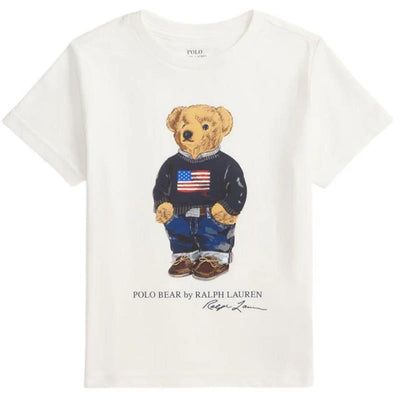 White short-sleeve t-shirt featuring a playful bear in a navy sweater with an American flag, styled with jeans and shoes.