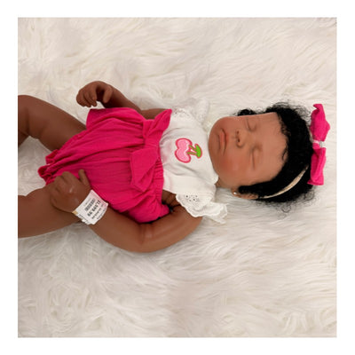 Reborn baby doll Emily, featuring closed eyes, African American skin tone, curly black hair, and a pink dress with cherry design.