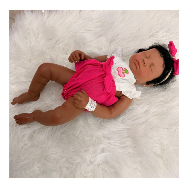 Reborn baby doll Emily, African American, with closed eyes, wearing a pink outfit and a bow, resting on a fluffy white surface.