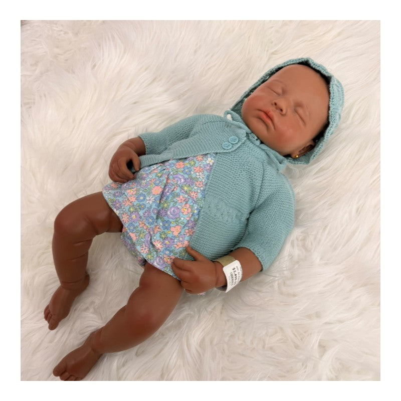 Reborn baby doll named Emily, with brown skin, wearing a blue knitted sweater and floral dress, resting on a white fluffy surface.