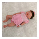 Reborn Baby Doll Vivienne, with a full silicone body, wearing a pink dress and headband, lying on a fluffy white surface.