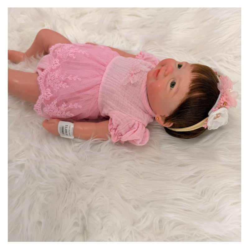 Reborn baby doll Vivienne in a pink dress with a floral headband, resting on soft white fabric.