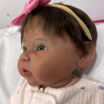 Reborn baby doll featuring white vinyl, dark brown hair, blue eyes, and a pink bow headband.
