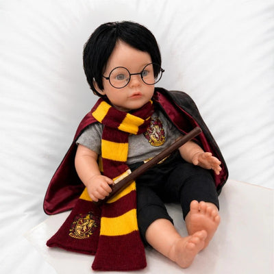 Reborn baby doll dressed as Harry Potter, featuring black hair, glasses, a Gryffindor scarf, and holding a wand.