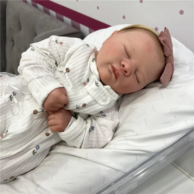 Reborn baby doll with white vinyl skin, wearing a floral onesie and a pink bow headband, sleeping on a soft surface.