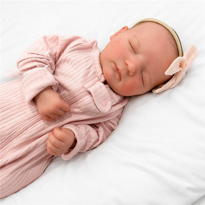 Reborn baby doll Jennie Asleep 2, with a pink ribbed onesie and a headband featuring a bow, resting on a white surface.