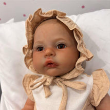 Reborn baby doll with brown eyes and light vinyl skin, wearing a ruffled bonnet and a white outfit, resting on a pillow.