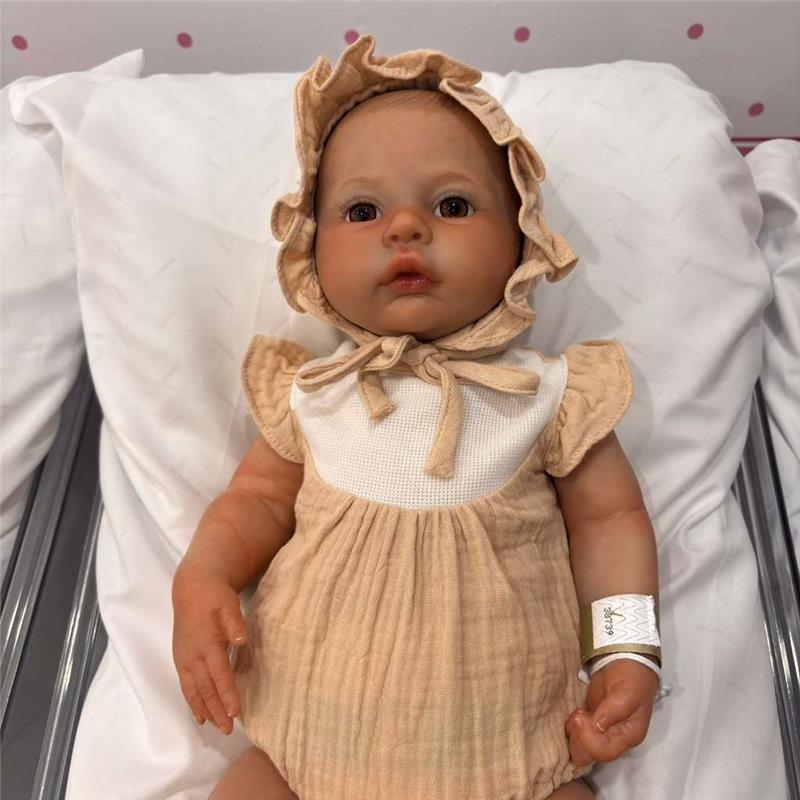 Reborn Baby Doll named Jocy, featuring a soft tan outfit, a matching bonnet, and realistic facial details, resting on a white blanket.