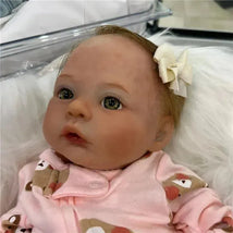 Reborn baby doll with light brown hair and green eyes, wearing a pink outfit with animal prints, adorned with a bow.
