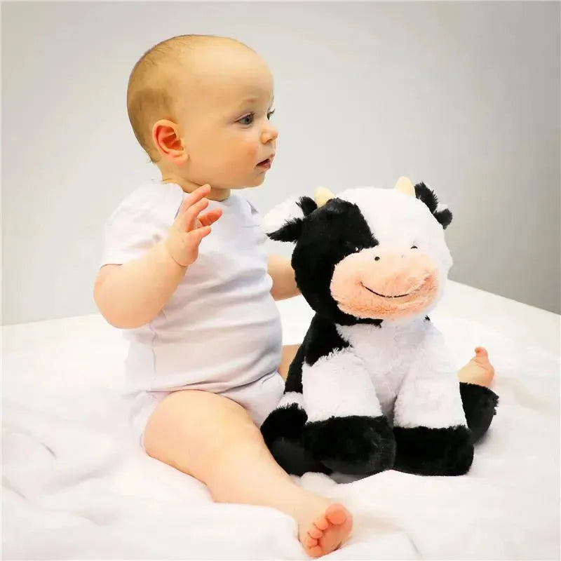 Baby in a white onesie sitting on a bed, next to a smiling plush cow toy.