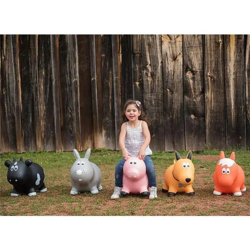 A young girl sits on a pink pig inflatable toy among other animal-shaped toys on grass, with a wooden backdrop.