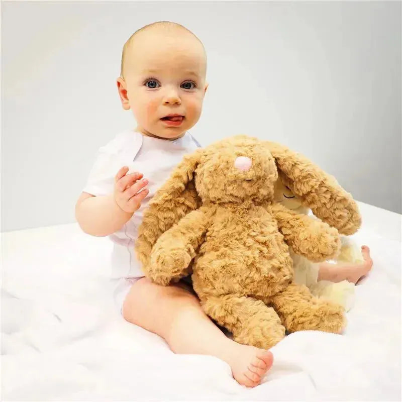Infant in a white outfit sitting on a soft surface, holding a textured plush bunny toy with floppy ears.