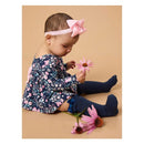 Baby girl wearing a floral navy dress and lace ruffle knee-high socks, sitting among pink flowers, looking at one.