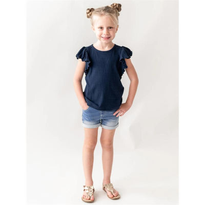 Light wash denim shorts on a smiling girl with a navy ruffled shirt, standing with hands on hips against a neutral background.